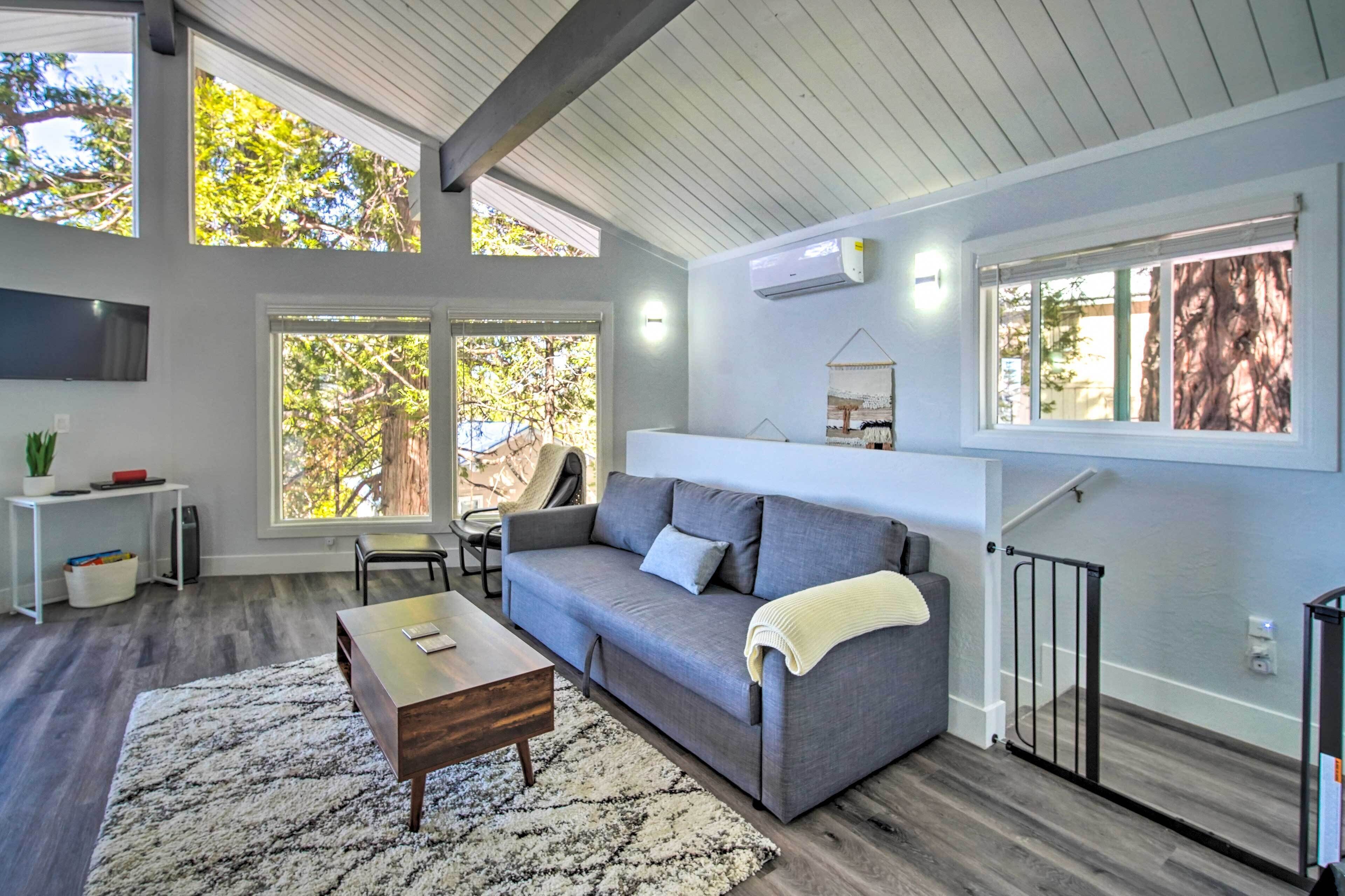 A modern living room with a grey sofa, a wooden coffee table, and a white rug. There are large windows allowing natural light and views of trees.