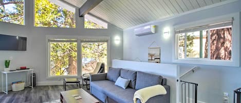 A modern living room with a grey sofa, a wooden coffee table, and a white rug. There are large windows allowing natural light and views of trees.