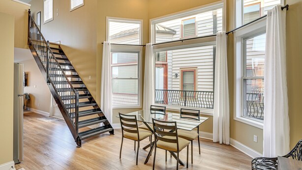 A dining area with a table and chairs, a staircase, and large windows with curtains.