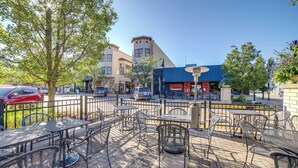 An outdoor patio with metal tables and chairs, a tree, and a building with a blue awning in the background.