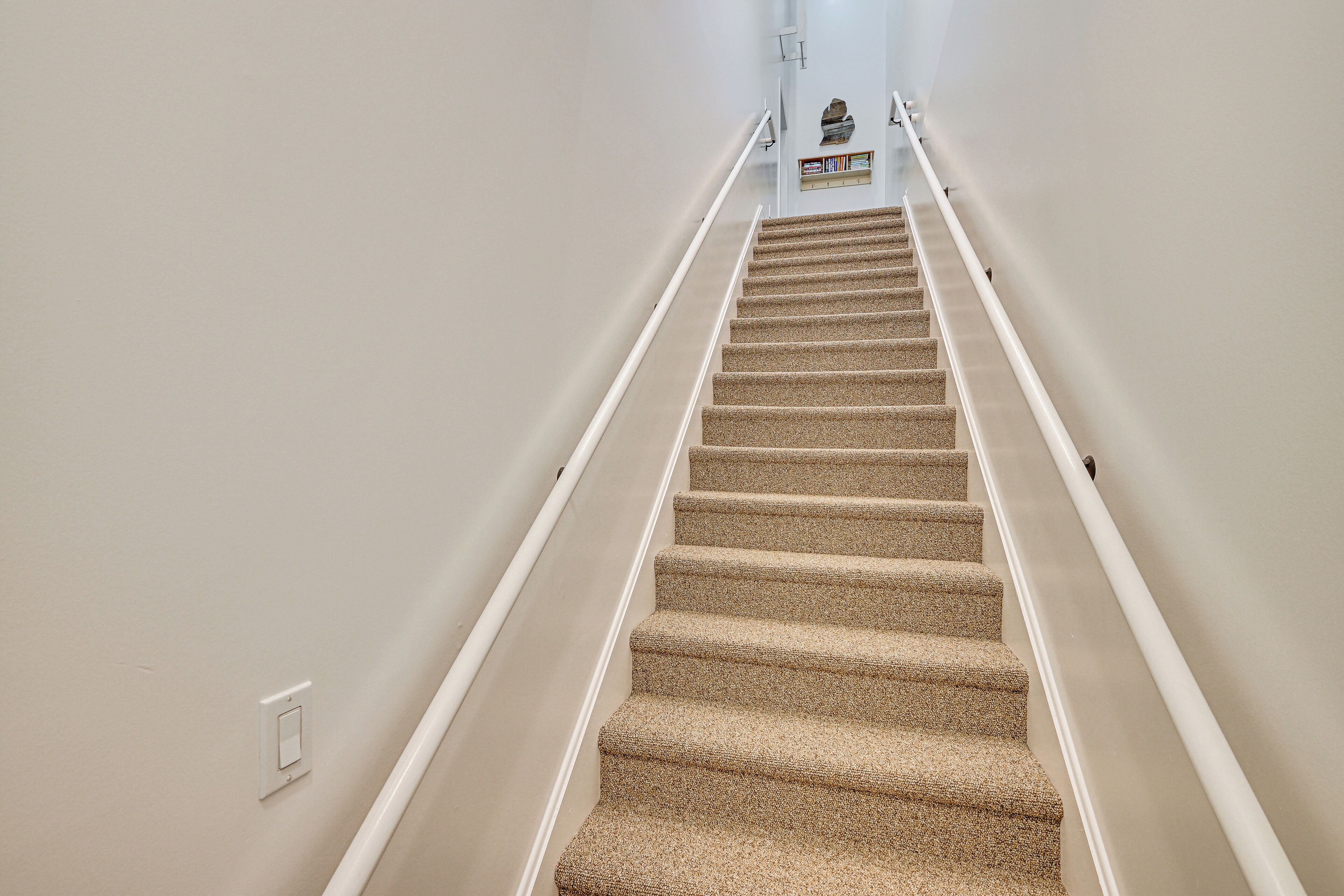 Eine Teppichstufen-Treppe mit weißen Geländern, die zu einer Zwischenebene mit einem kleinen Regal und einem dekorativen Gegenstand führt.