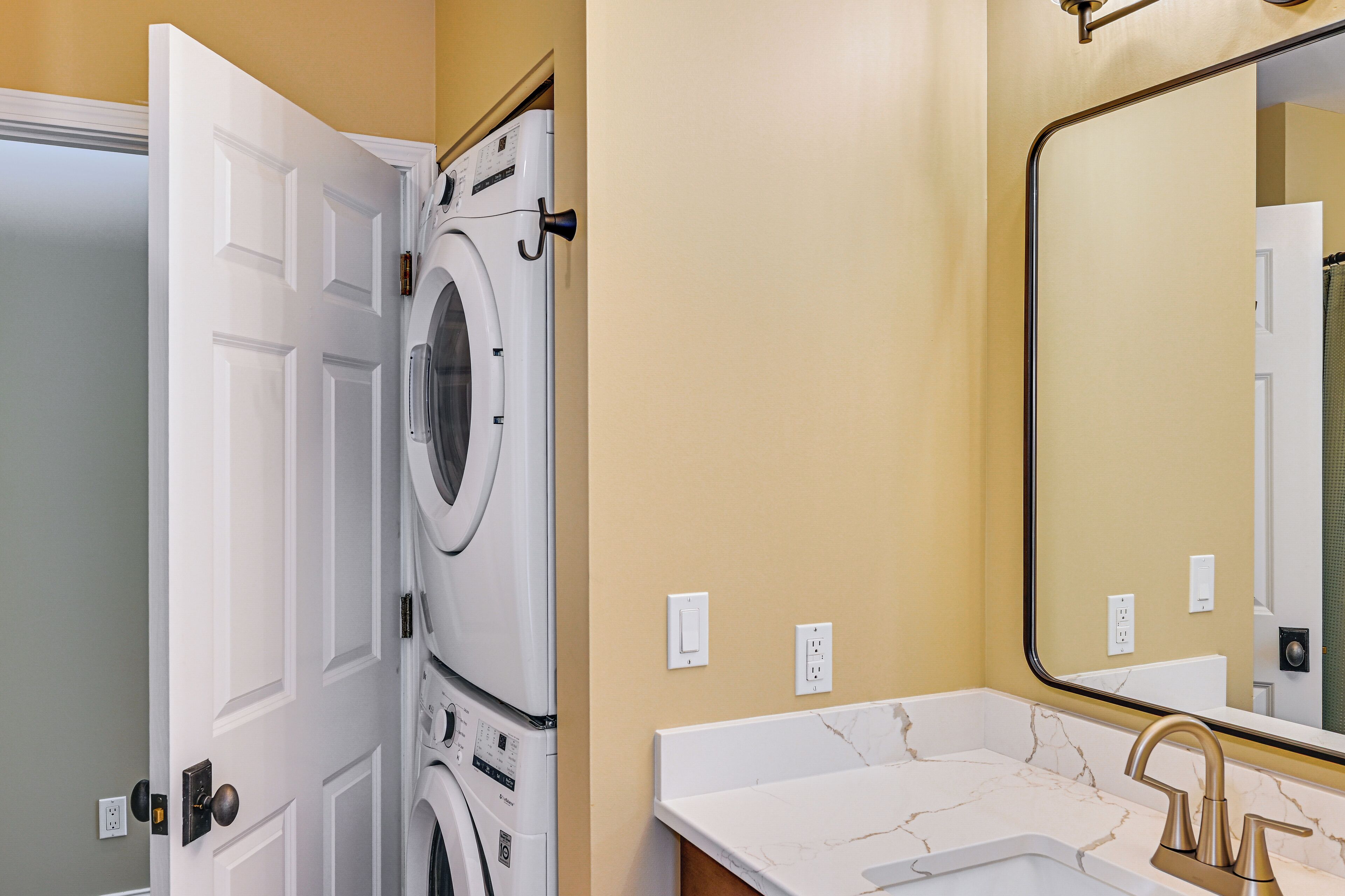 A laundry room with stacked washing machines, a white door, and a bathroom with a sink and mirror.