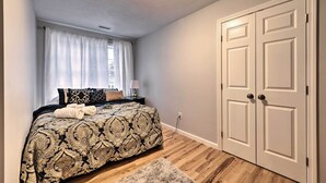 A bedroom with a bed, a nightstand, a lamp, a window with curtains, a door, and a wooden floor. - Worcester Vacation Rental w/ Ideal Location! (Worcester)