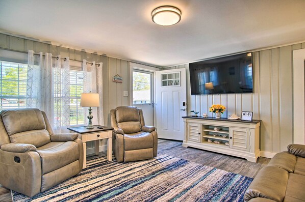 A living room with a beige leather recliner, a small table with a lamp, a TV on a white cabinet, and a striped rug.