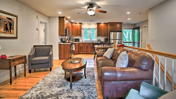 A spacious living room with a leather sofa, a wooden coffee table, and a chair. A kitchen with wooden cabinets and a large window is visible in the background. - Cape Cod Home w/ Deck ~ 3 Mi to Beaches (Buzzards Bay)