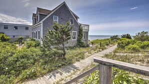 A coastal house with a wooden deck leading to a sandy beach. - Fireplace + Sunroom! Oceanfront Cape Cod Cottage (Dennis Port)