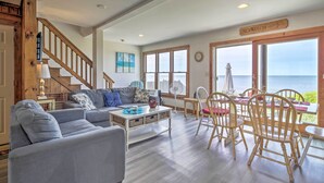 A spacious living area with a staircase, a dining table with chairs, and large windows overlooking the sea. - Fireplace + Sunroom! Oceanfront Cape Cod Cottage (Dennis Port)