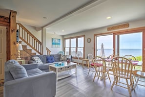 A spacious living room with a large window overlooking the sea, a wooden staircase, and a dining area with wooden chairs and a table. - Fireplace + Sunroom! Oceanfront Cape Cod Cottage (Dennis Port)