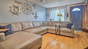 Una sala de estar con un sofá seccional color beige, un ventilador de techo, decoración de pared con temática náutica, una mesita auxiliar y piso de madera.