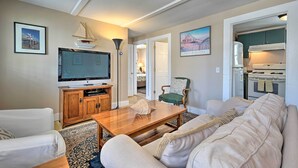 Un salon avec une télévision, une table basse en bois et un canapé. On peut apercevoir une cuisine à travers une porte ouverte.