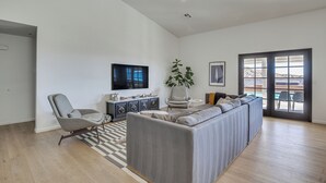 Uma sala de estar ampla com um sofá seccional grande, uma poltrona cinza, uma TV de tela plana, um vaso de planta e vista para o exterior através de uma porta de vidro.