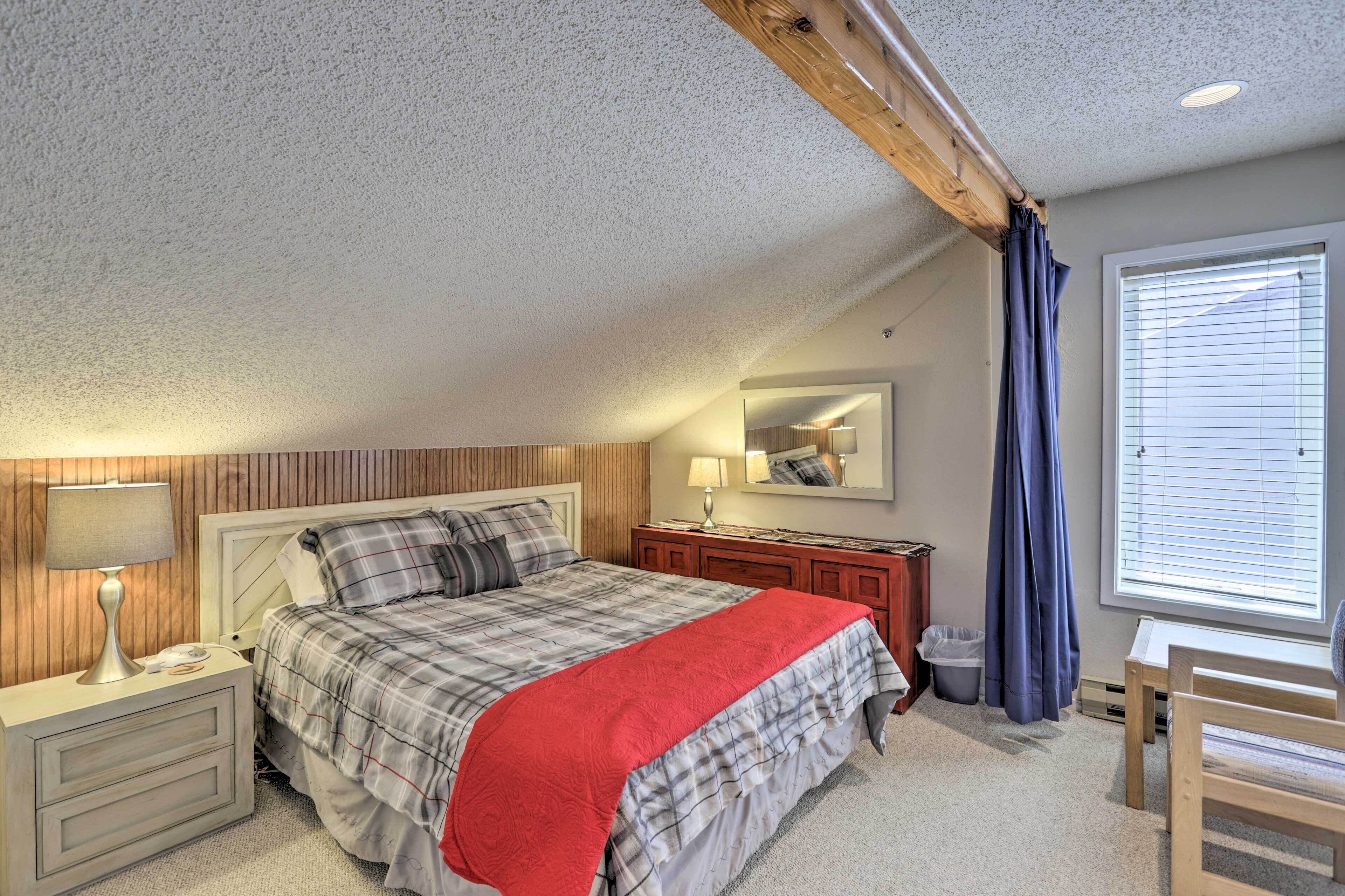 A bedroom with a bed, nightstands, a dresser, a mirror, a window with blinds, and a wooden ceiling beam.