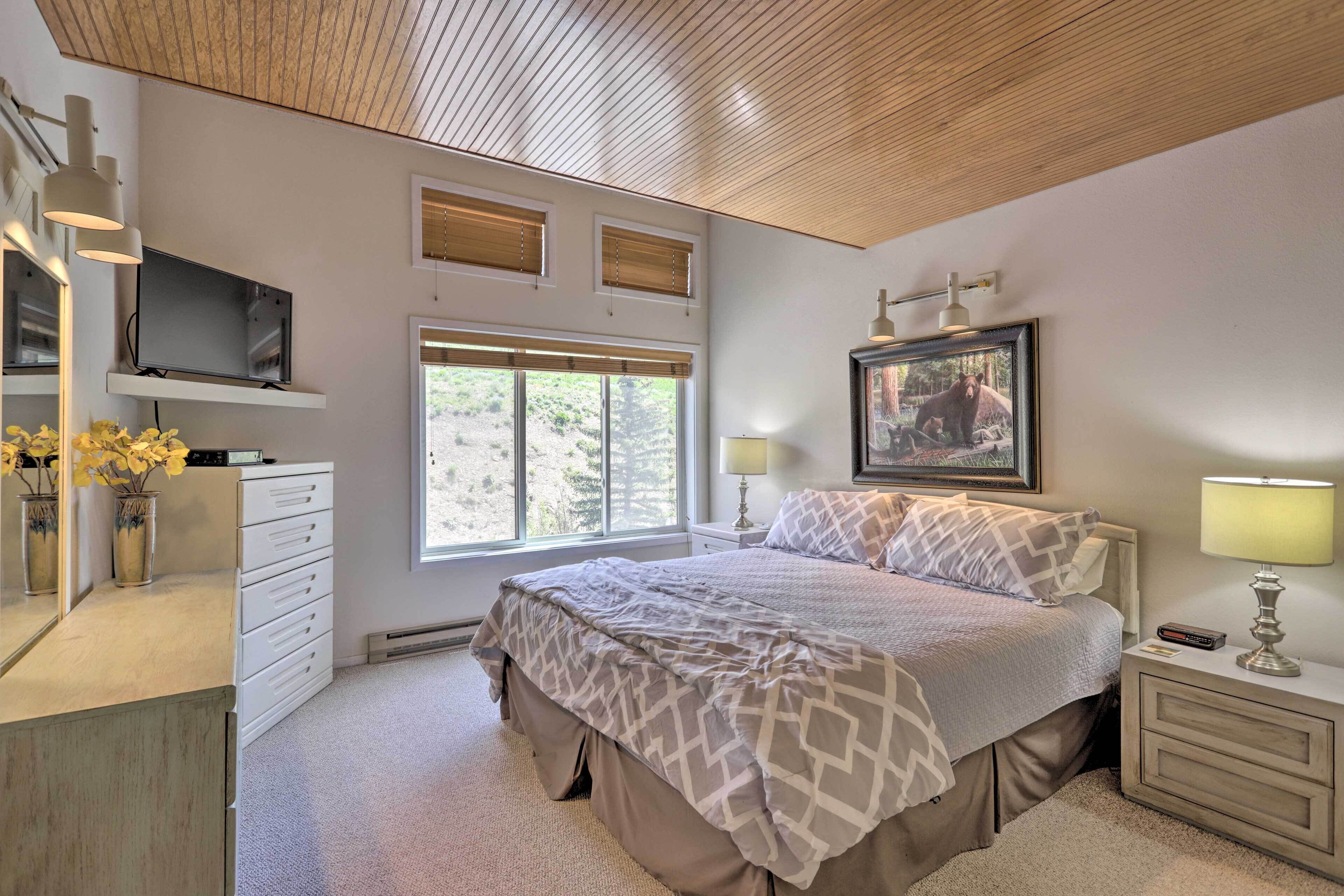 A bedroom with a bed, bedside tables, a TV, a window with blinds, a painting, and a mirror.