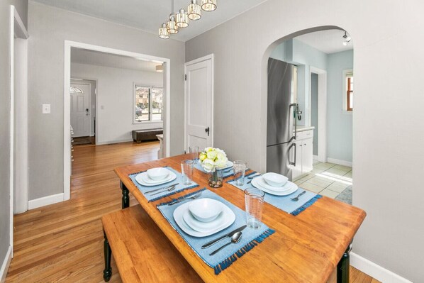 A dining area with a wooden table set for four, a refrigerator, and a bench in the background.