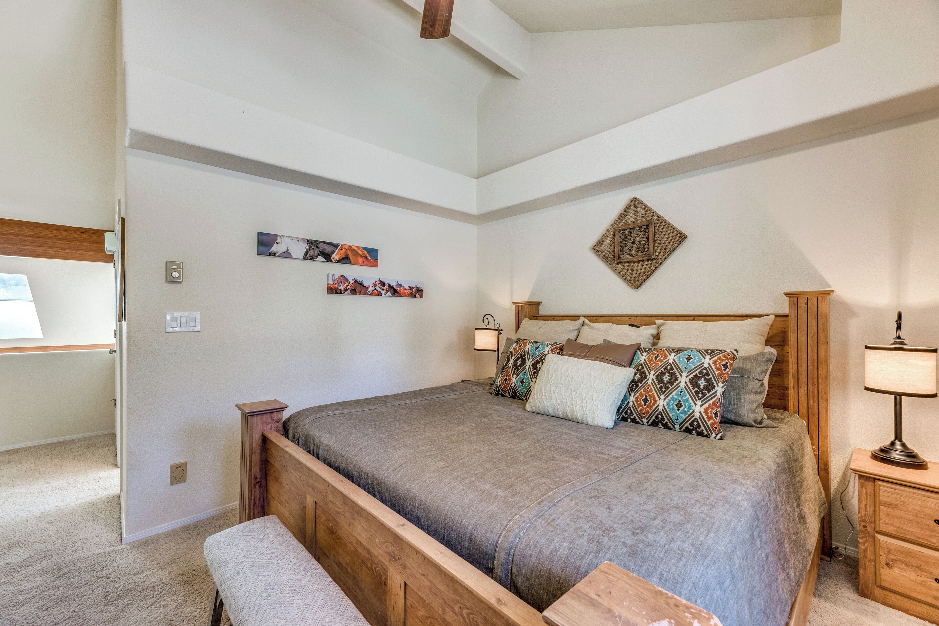A bedroom with a large bed, wooden headboard, and a bench. There are wall-mounted lights and a small table with a lamp.