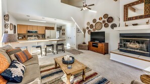 Amplia sala de estar con sofá, mesa de centro y chimenea. Se observa una cocina al fondo con armarios de madera y un comedor.