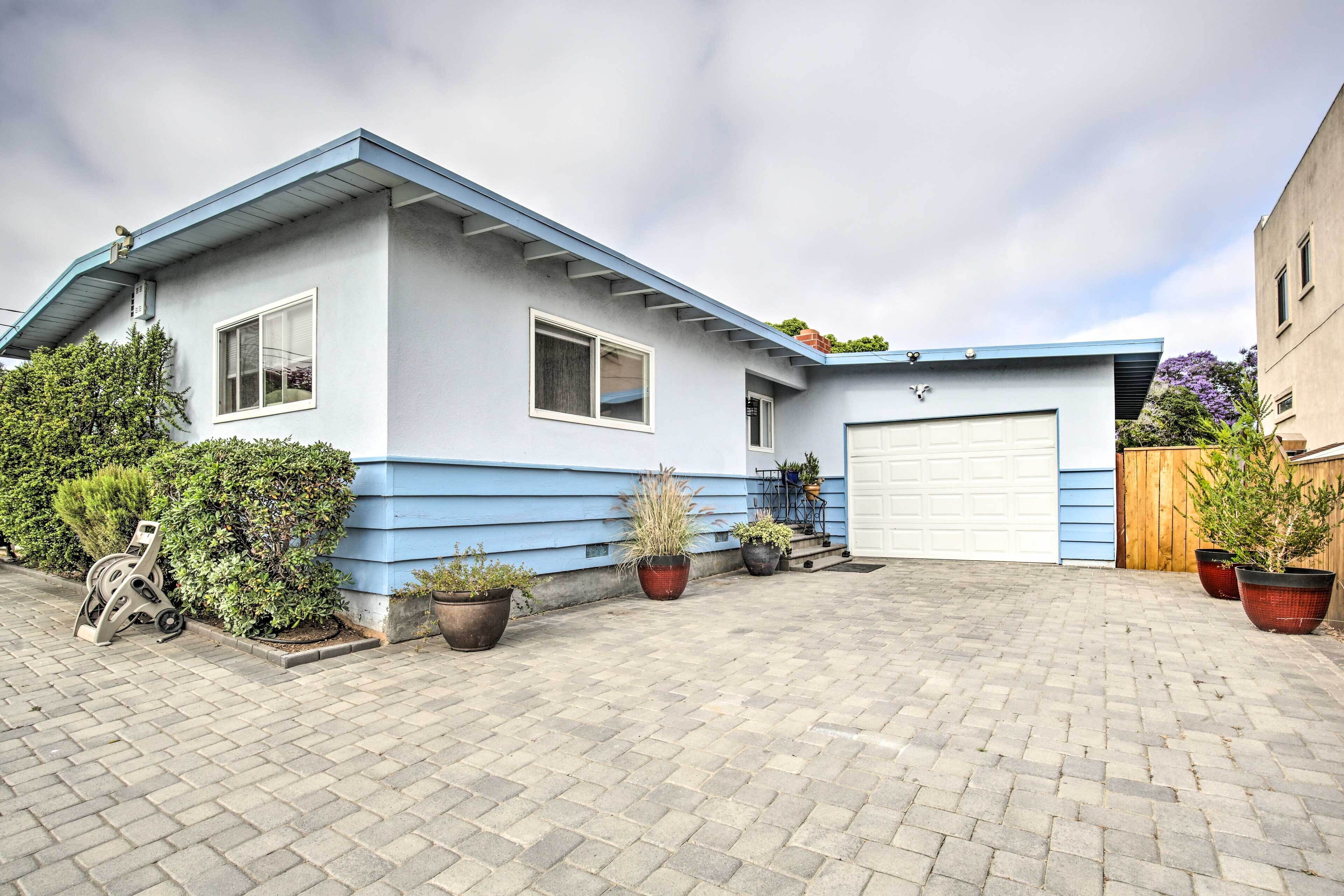 A two-story house with a blue exterior and a garage. There are potted plants along the driveway and a stroller parked outside.