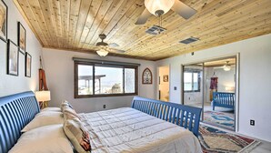 A bedroom with a wooden ceiling, a large bed with a blue headboard, a window with a view, and a mirror reflecting the room. - Chic Joshua Tree Pad w/ Mountain Views & Hot Tub (Twentynine Palms)