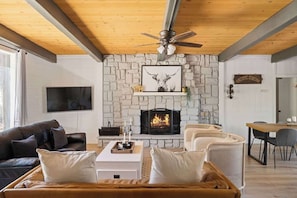 Un salon doté d’une cheminée en pierre, d’un ventilateur de plafond, d’un téléviseur à écran plat et d’un coin repas avec une table et des chaises en bois.