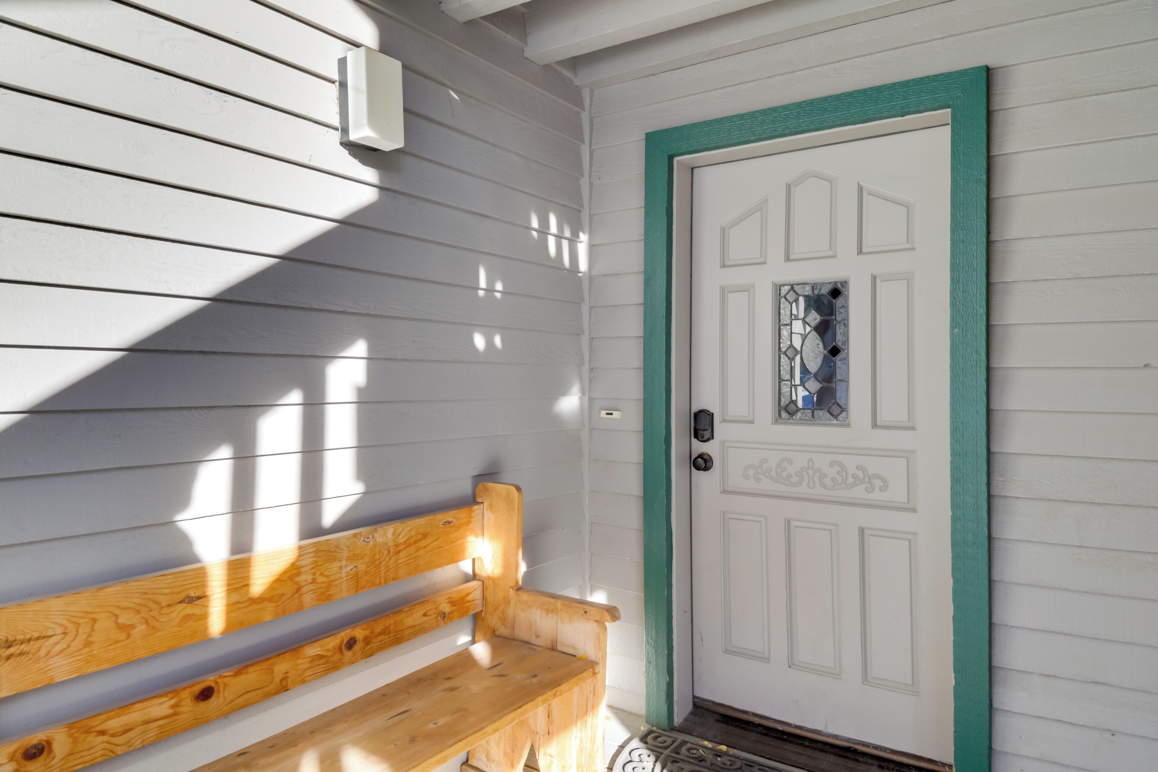 A wooden bench outside a white door with a green trim and a decorative glass panel.