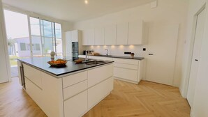 A modern kitchen with white cabinetry, a large island with a sink, and a view of the outside. - Luxury villa with swimming pool for 8 people (LA COUARDE SUR MER)