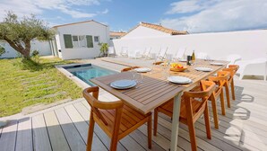 A wooden deck with a pool, outdoor dining set, and a white building with green shutters. - Luxury villa with swimming pool for 8 people (LA COUARDE SUR MER)