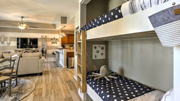 Una cama litera con una colcha de lunares, una silla de madera, un televisor y una cocina al fondo.