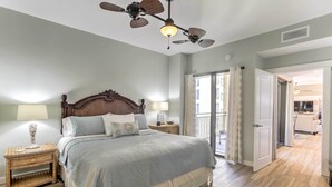 Un dormitorio con una cama grande, dos mesitas de noche con lámparas, un ventilador de techo y vistas al salón a través de una puerta abierta.