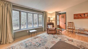 A cozy living room with a large window, a brown armchair, a wooden chair, a bench, and a patterned rug. - Four-season Home on Scenic Bluff Country Acreage! (Black River Falls)