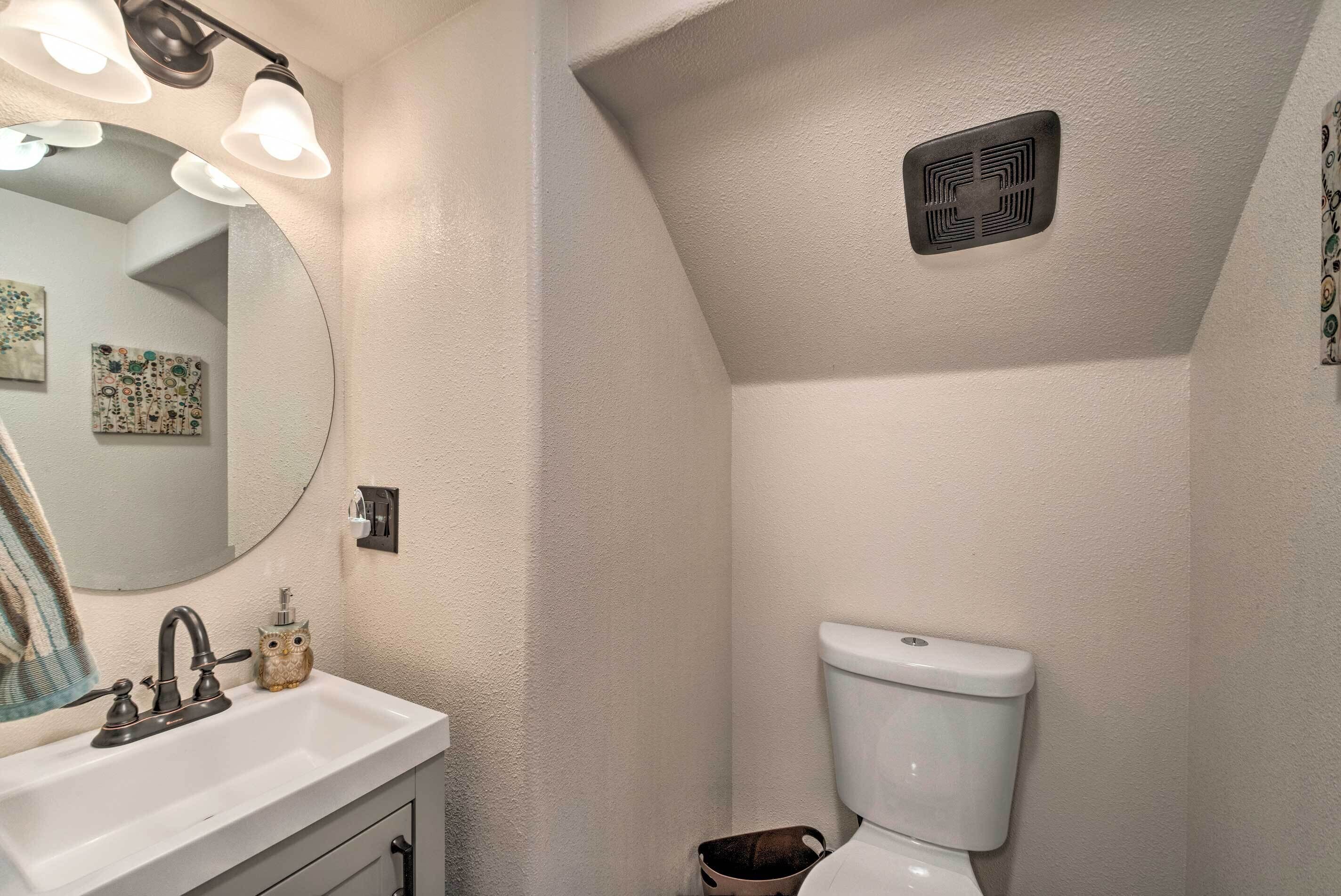 Un baño con un lavamanos blanco, un espejo, un inodoro y una rejilla de ventilación en el techo.