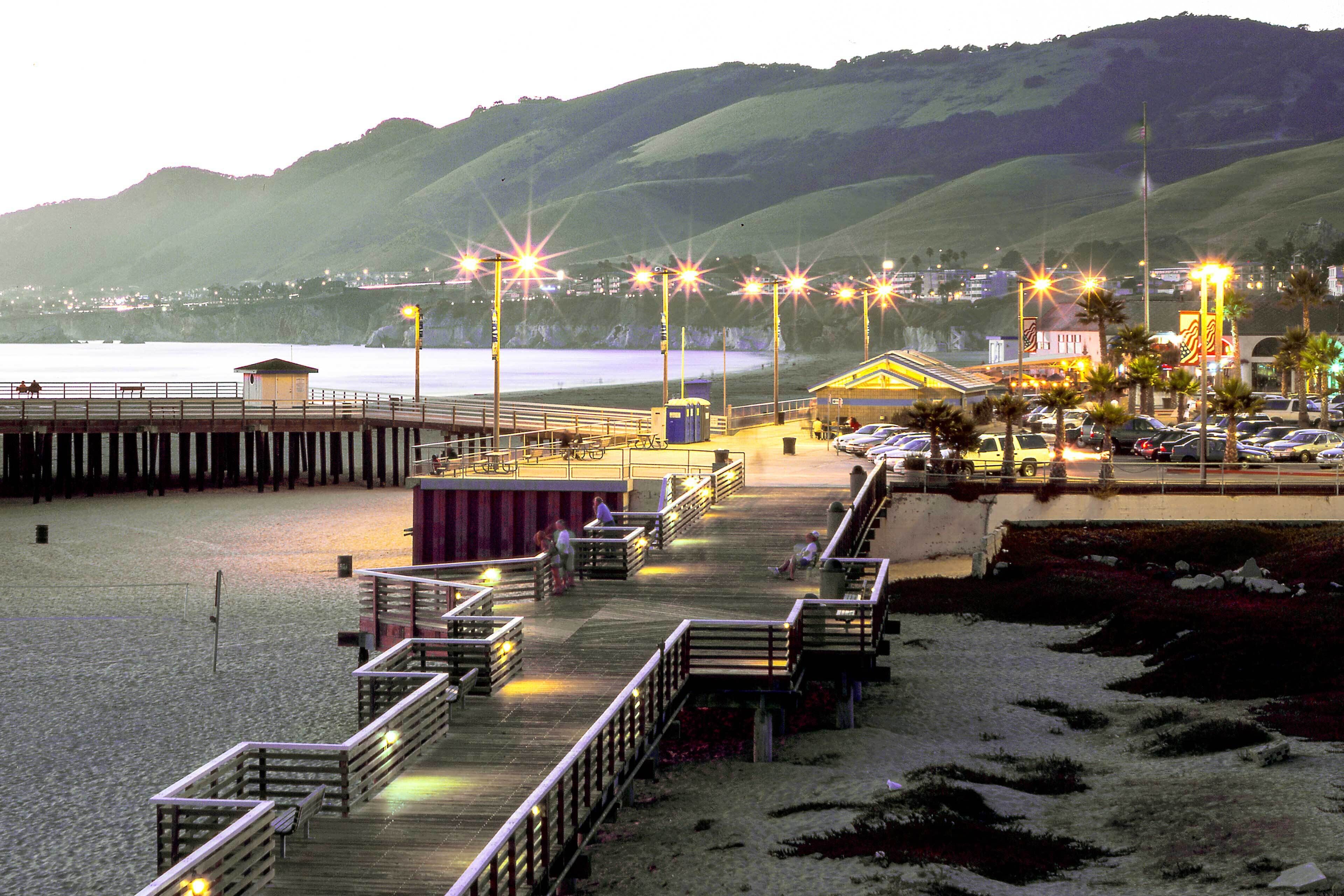 A pier with a wooden walkway leading to a building with a yellow roof, overlooking a beach and hills.