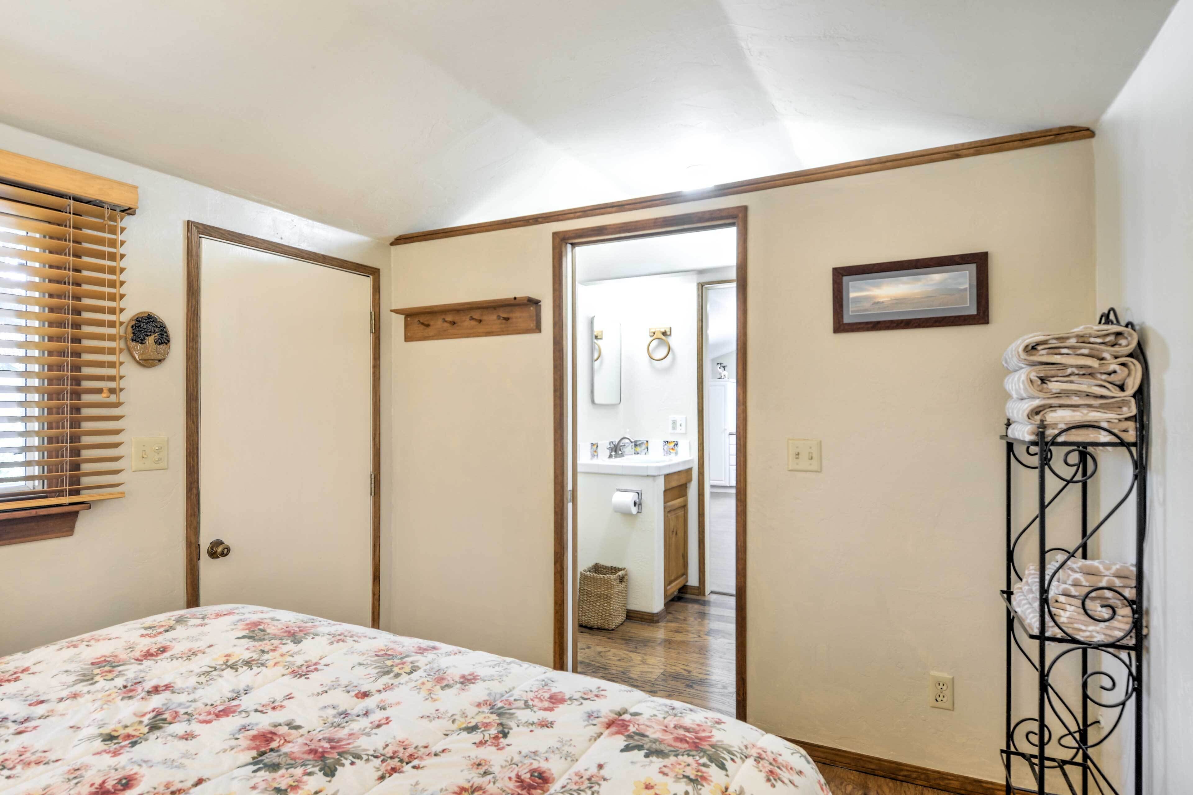 Quarto com colcha florida, porta de madeira, espelho, porta-toalhas e vista para o banheiro.