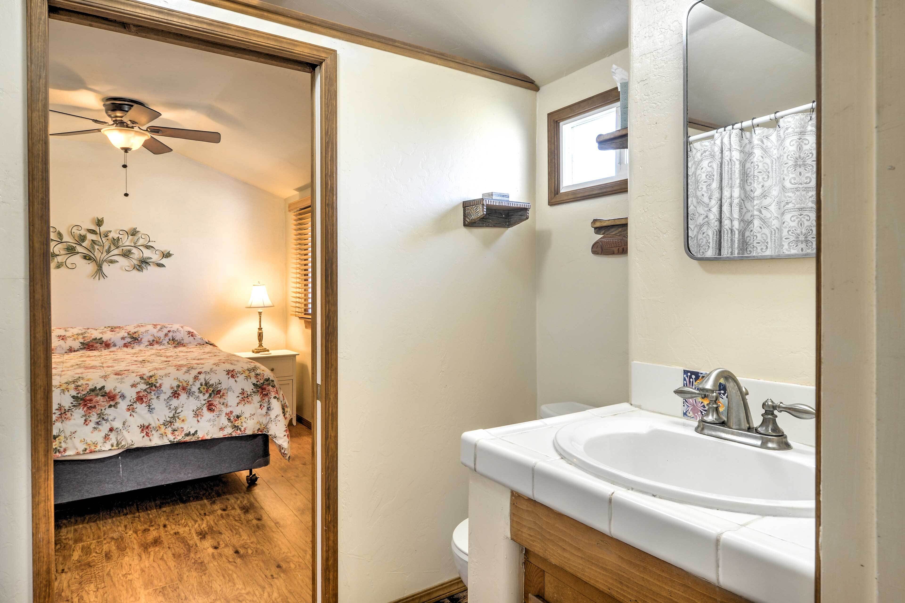 A bedroom with a bed, a ceiling fan, a nightstand with a lamp, and a bathroom with a sink and mirror.