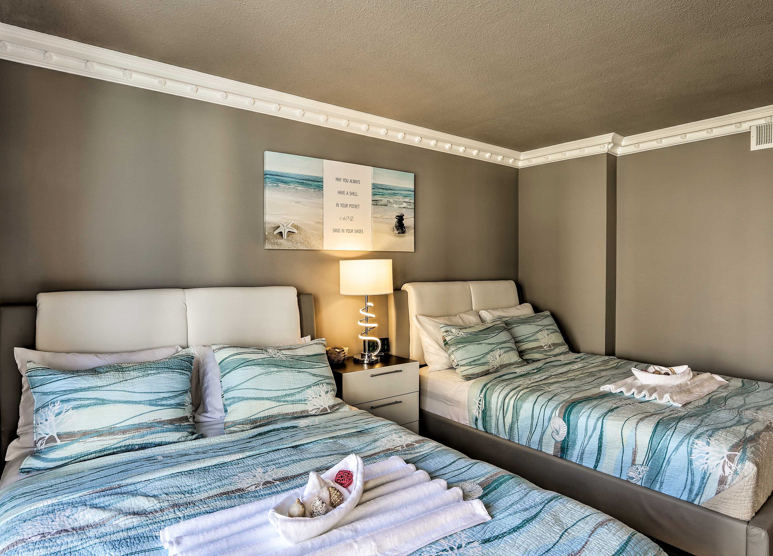 A hotel room with two beds, each with a striped blue and white bedding. There is a wall-mounted artwork with a beach scene and text, a bedside table with a lamp, and a small decorative item on the nightstand.