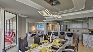 A modern dining area with a long table set for six, a kitchen with grey cabinets, and a view of a balcony with red chairs.