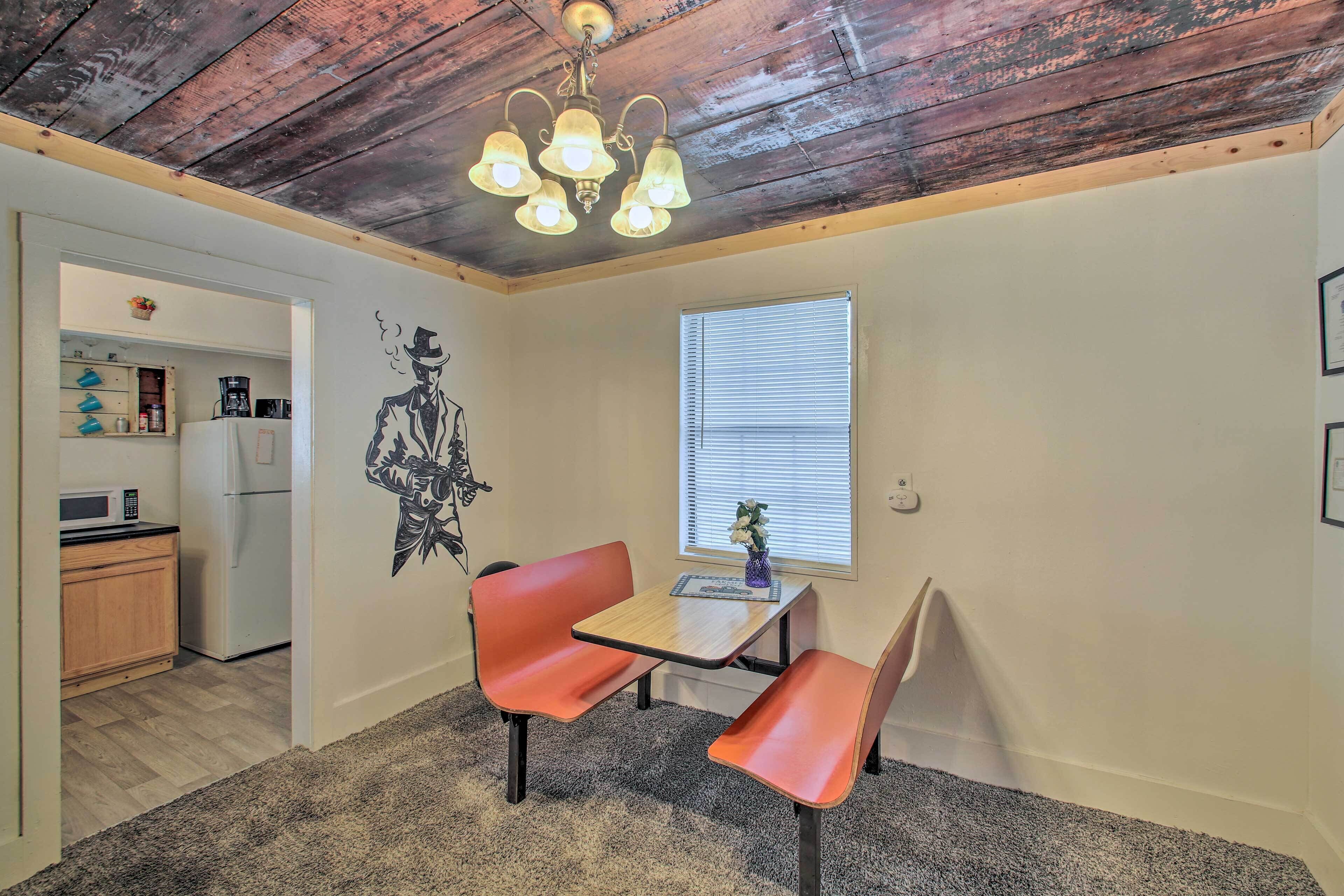 A dining area with a table and two chairs, a ceiling light fixture, a window with blinds, and a kitchen in the background.
