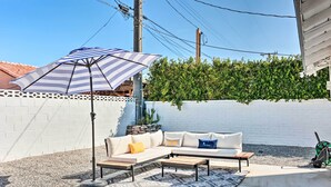 A patio with a white brick wall, a white fence, a green hedge, a striped umbrella, a white sofa with cushions, and a patterned rug. - < 4 Mi to Dtwn Palm Springs: Home w/ Private Patio (Palm Springs)