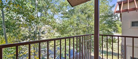 A wooden balcony with a metal railing overlooking a landscaped area with trees and a building in the background.