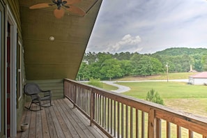 Une terrasse en bois avec une chaise berçante et un ventilateur de plafond, d’où l’on peut admirer un paysage verdoyant, avec une route sinueuse et une colline au loin.