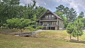 Une maison en bois à deux étages, dotée d’un balcon, entourée d’arbres et traversée par un petit pont.