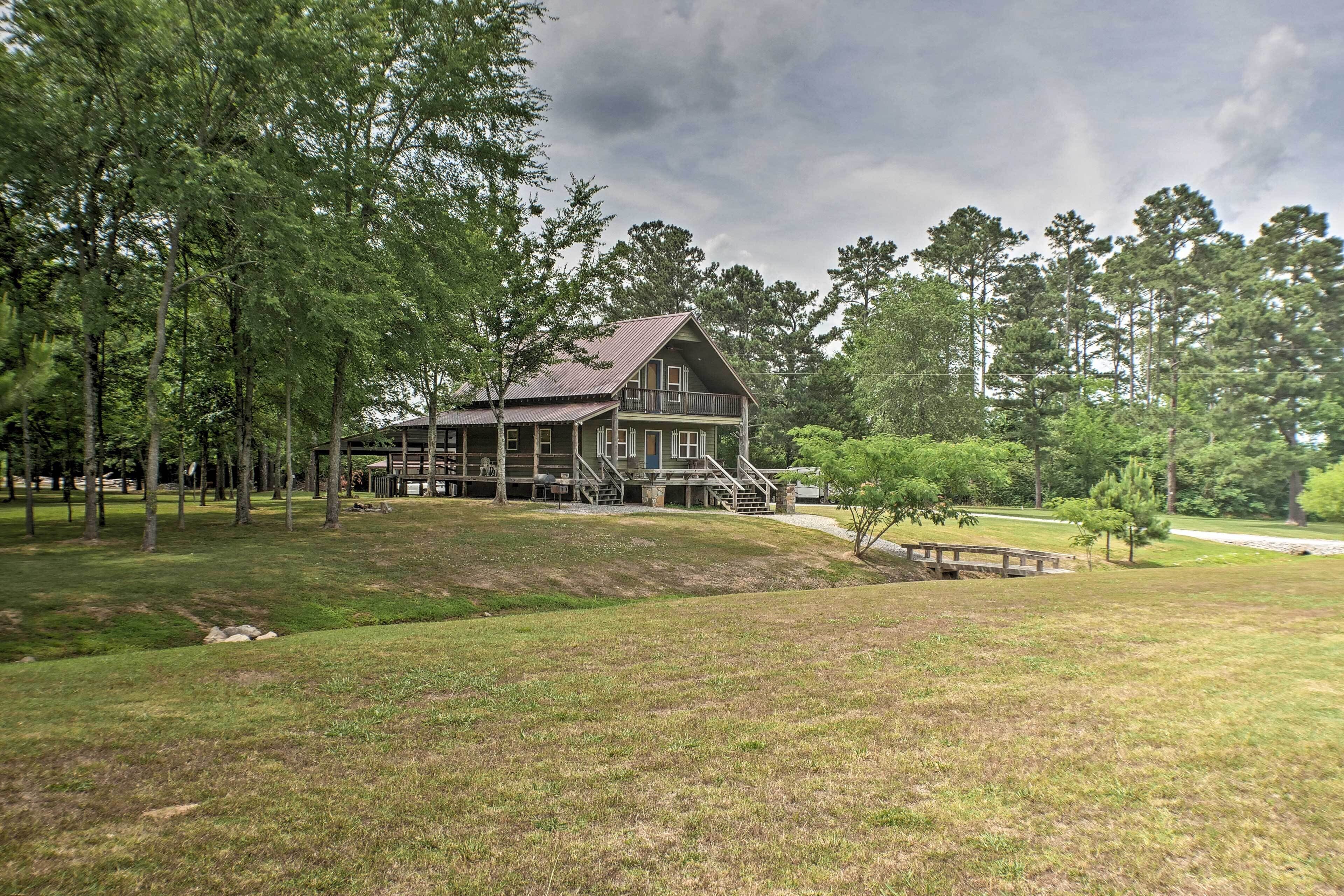 A large, two-story wooden house with a porch and a small bridge leading to it, surrounded by a grassy area and trees.