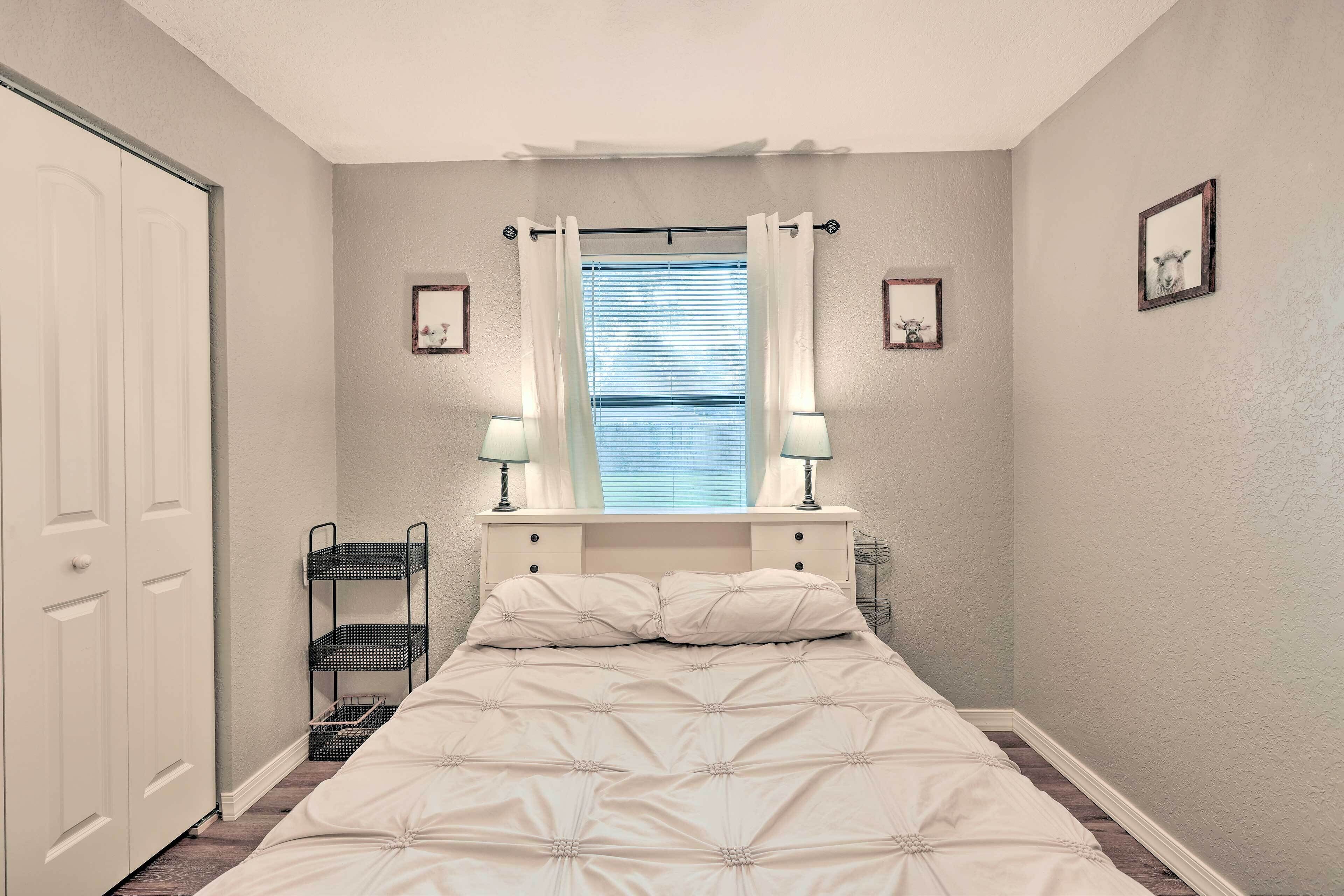 Un dormitorio con cama, ventana con cortinas, mesita de noche con lámparas y estante fijado a la pared.