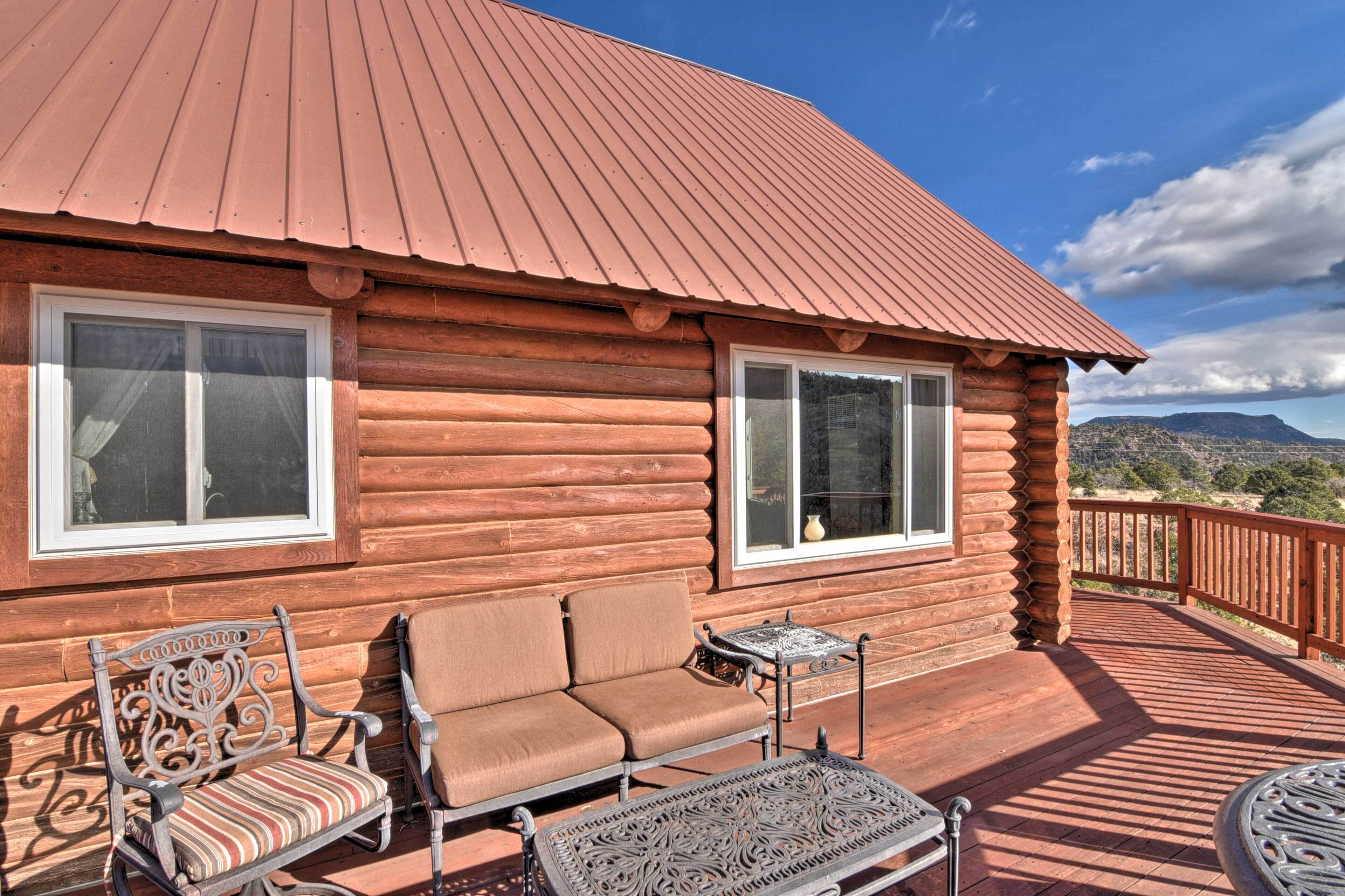 Une cabane en rondins avec un toit en métal, deux fenêtres, une terrasse avec du mobilier d’extérieur et une vue panoramique sur les montagnes.