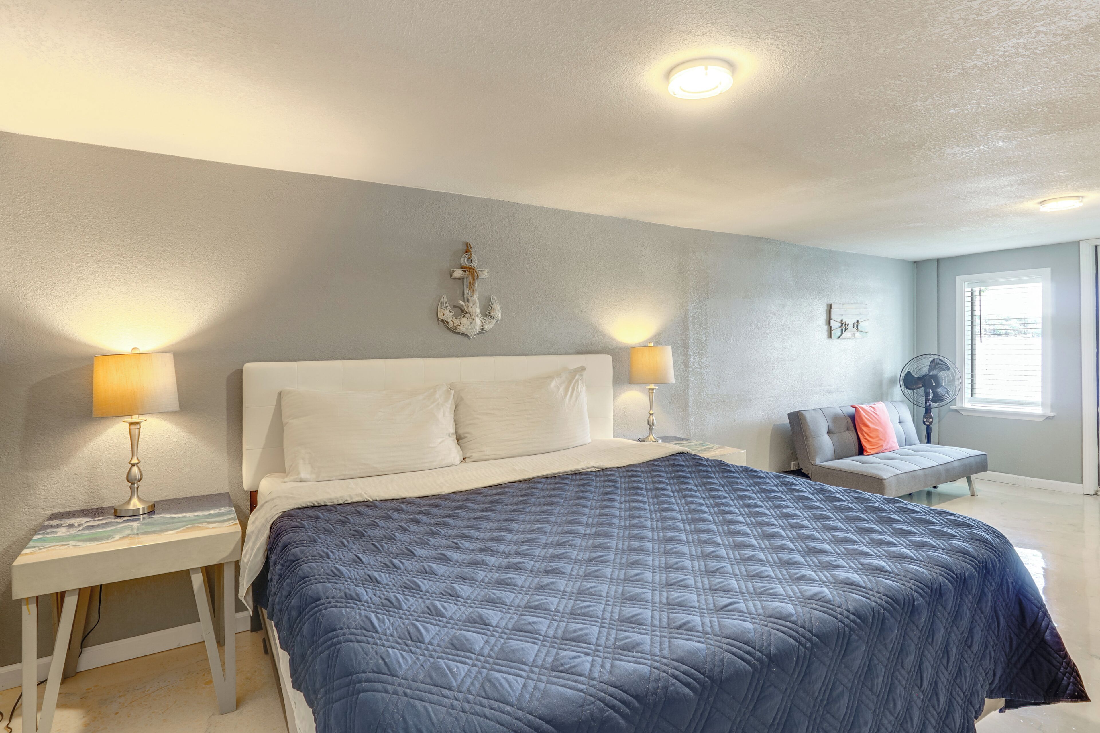 Ein Schlafzimmer mit einem großen Bett, zwei Nachttischen, einem Beistelltisch, einem Wandventilator und einem Fenster mit Jalousien.