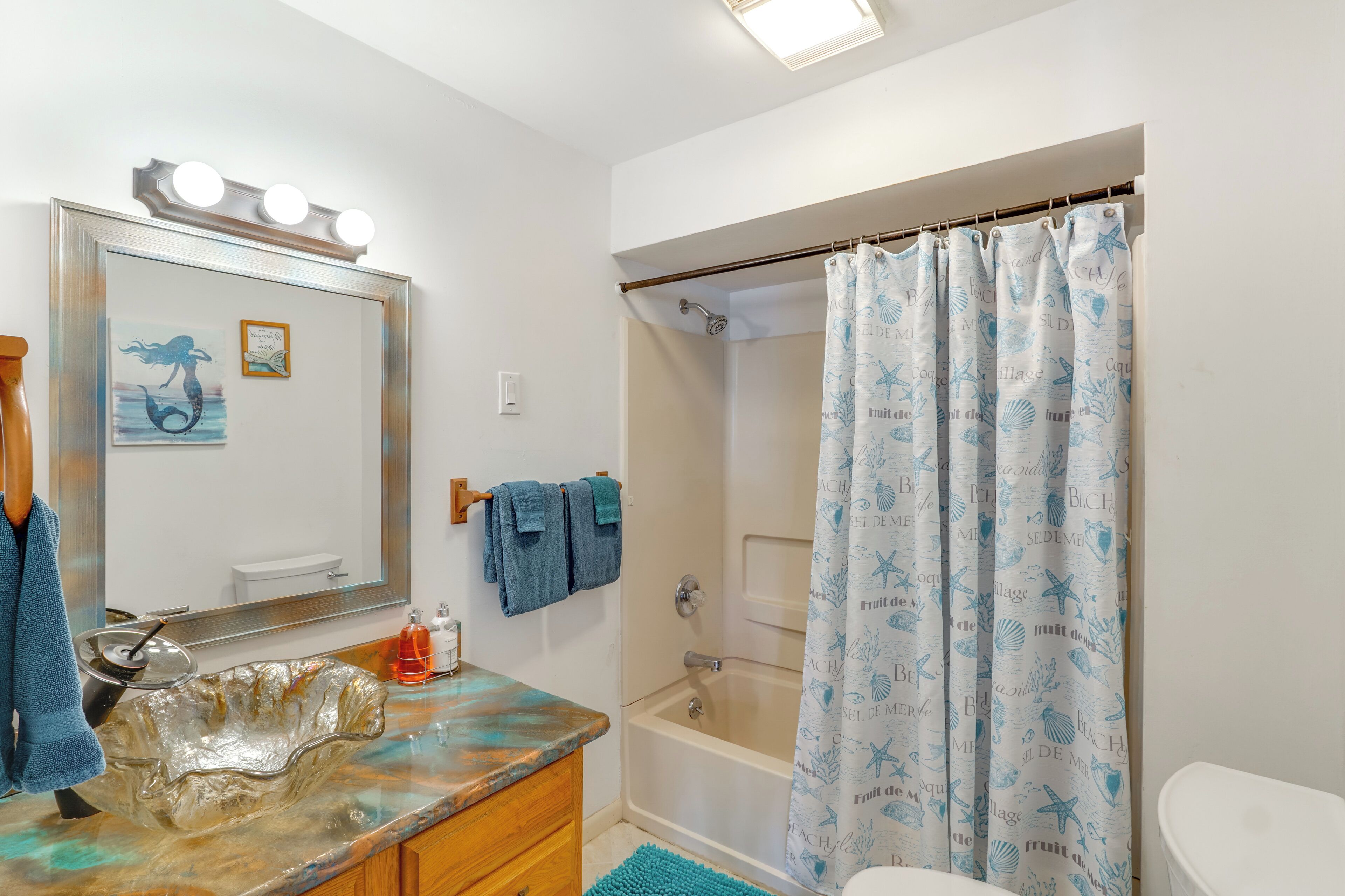 Ein Badezimmer mit einem Glaskonfett, einem beleuchteten Spiegel, einer Duschvorhang mit Strandmotiv und einer Toilette.
