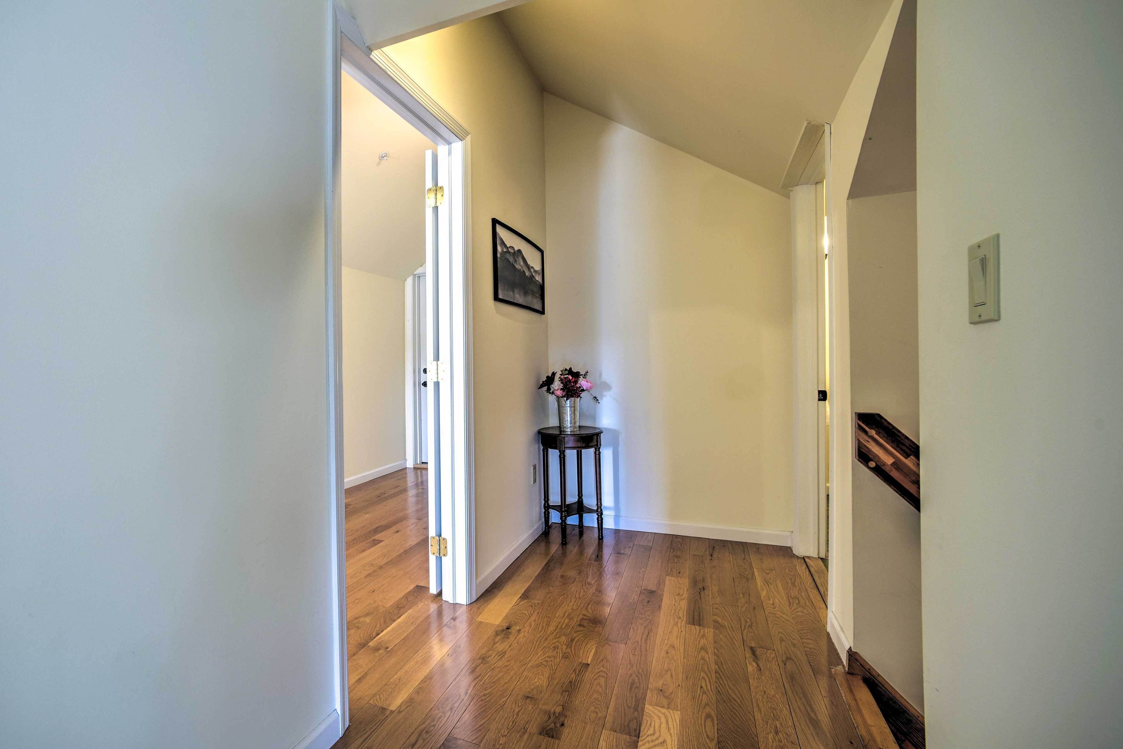 A hallway with wooden flooring, a door leading to another room, a small table with a vase of flowers, and a framed picture on the wall.
