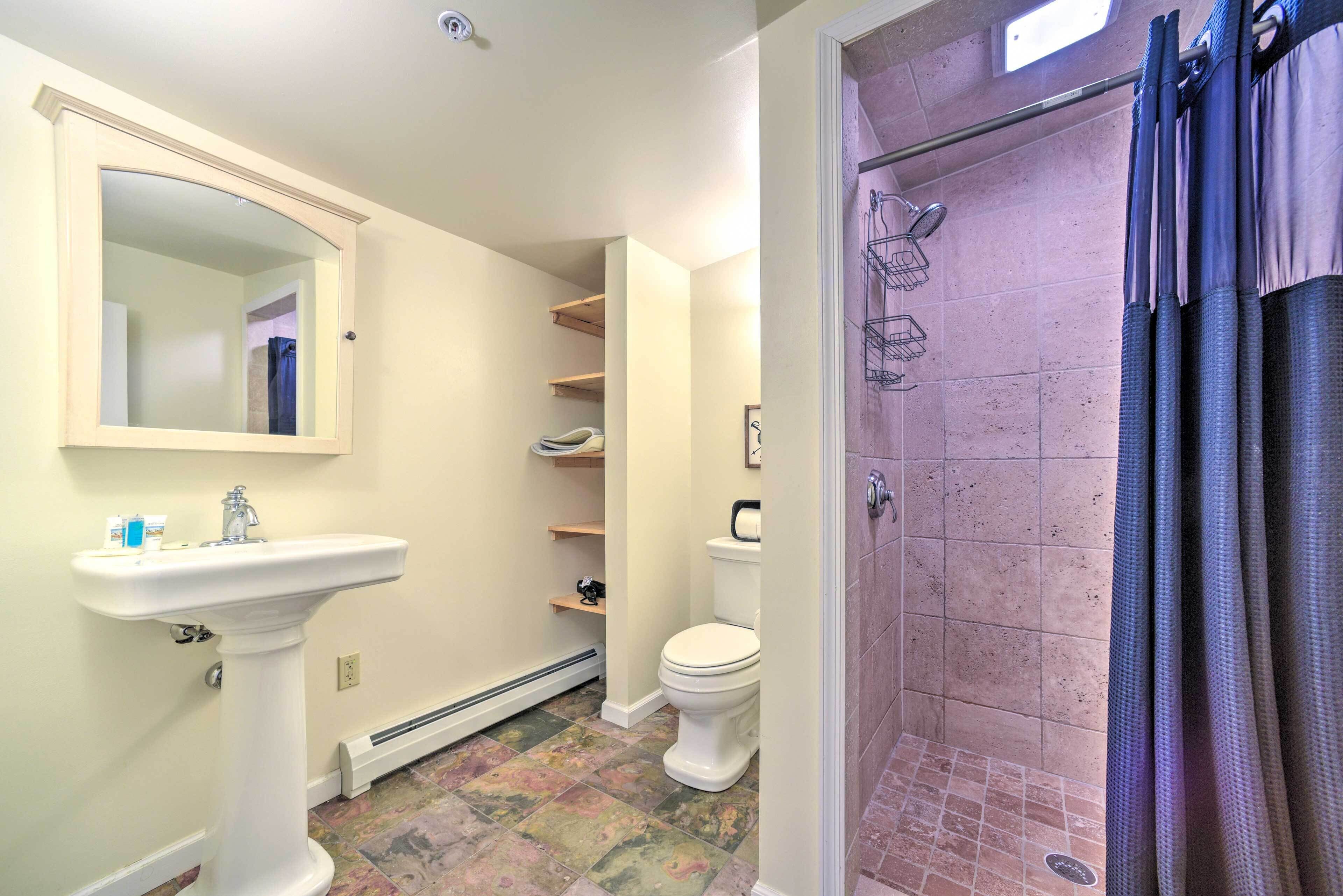 A bathroom with a pedestal sink, a toilet, a shower with a glass door, and a mirror. Shelves are mounted on the wall above the toilet.