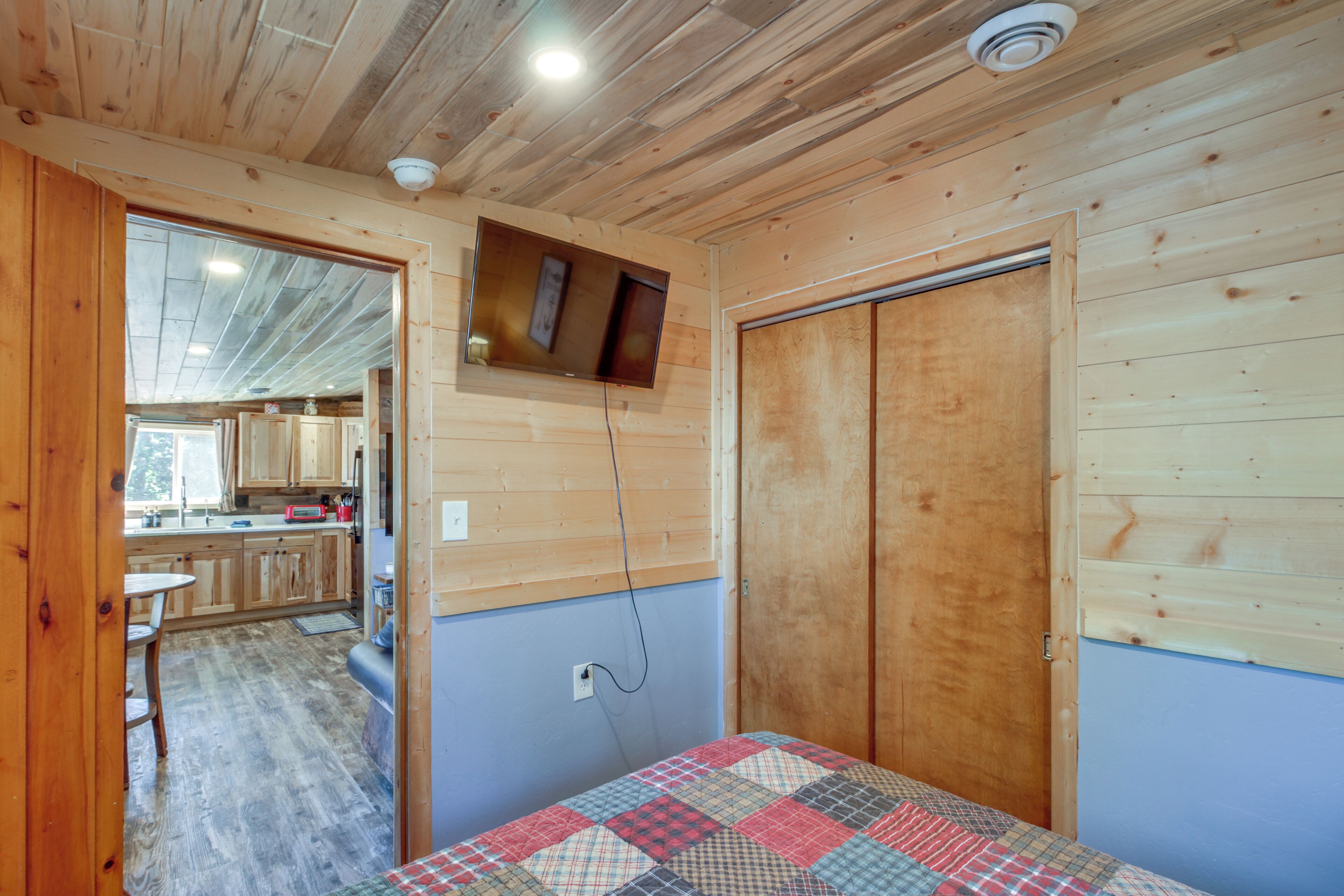 A wooden interior with a quilted bed, a wooden door, and a kitchen visible through an open doorway.