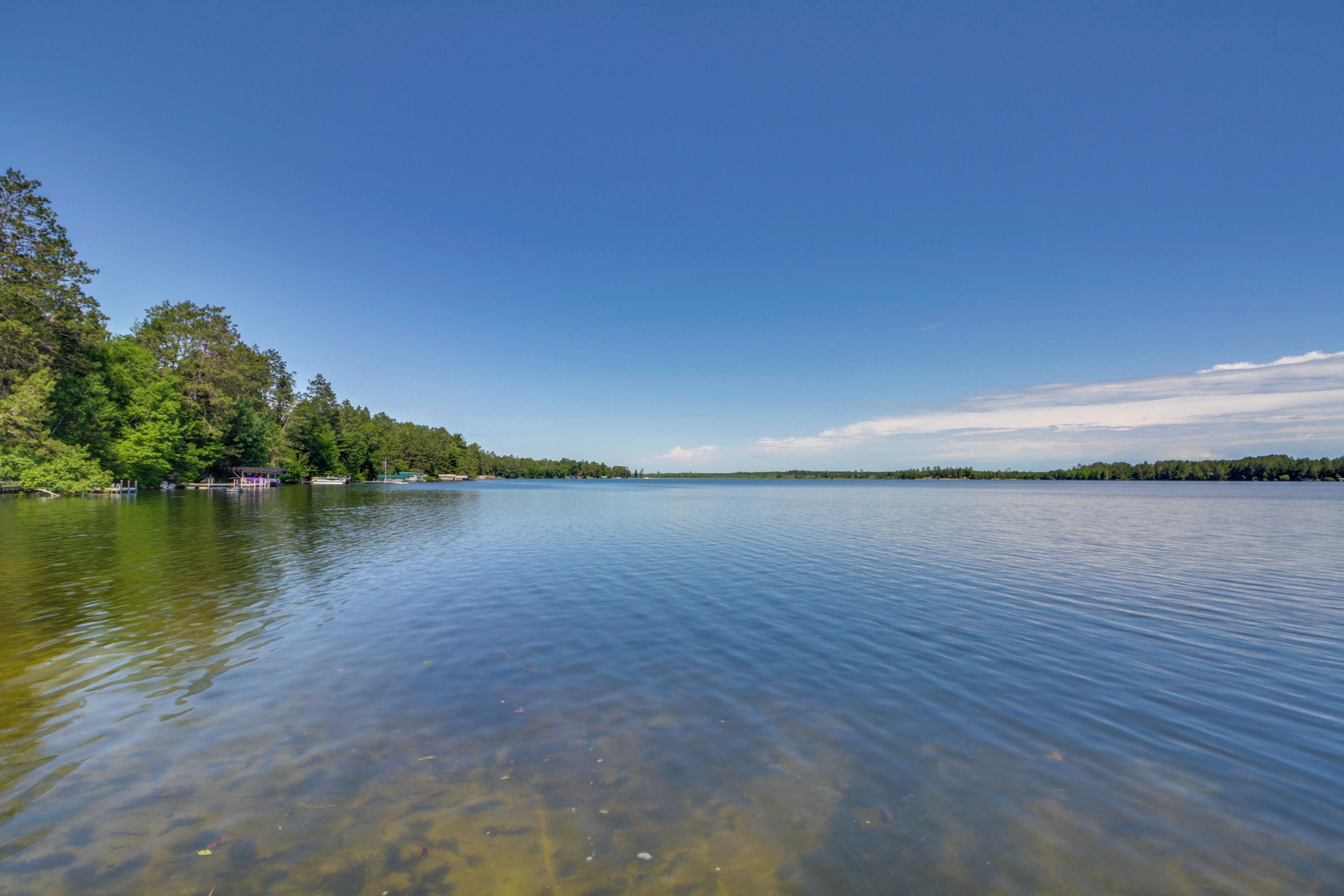 A calm lake with clear water, surrounded by dense green trees and a distant shoreline.