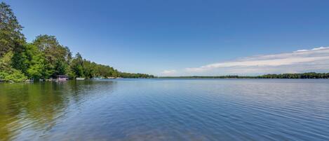 A calm lake with clear water, surrounded by dense green trees and a distant shoreline.